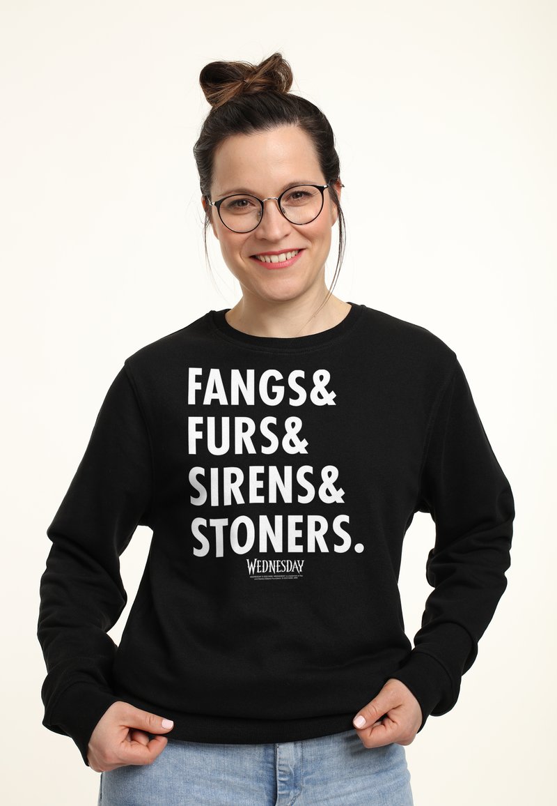 Persona sorridente con occhiali che indossa un felpa nera con la scritta "Fangs & Furs & Sirens & Stoners" in bianco, mani che afferrano l'orlo.