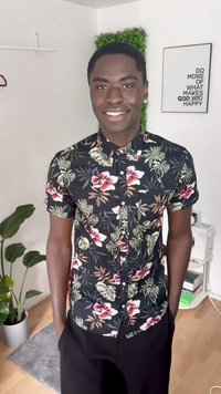 Camicetta a maniche corte con bottoni nera con motivi floreali in rosso, rosa e verde. Abbinata a pantaloni neri; il modello è in piedi con le mani in tasca.