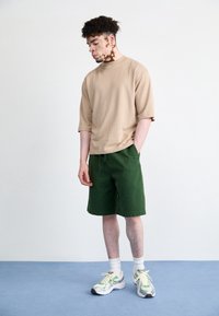 Camiseta de gran tamaño en color beige, pantalones cortos verdes, calcetines blancos y zapatillas multicolores. Mangas cortas y un ajuste relajado; acabado de tela suave.