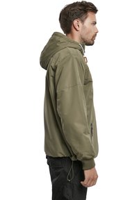 Veste à capuche vert olive avec une poche avant zippée, des poignets élastiques et un cordon de serrage réglable à l'ourlet. Texture légèrement rembourrée.