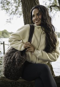 Sudadera con cremallera de forro polar beige con tejido texturizado, combinada con un bolso de hombro de felpa marrón oscuro. El modelo está sentado sobre un tocón de árbol junto al agua.