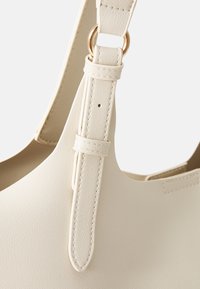 Beige käsilaukku, jossa on sileä, teksturoidulla pinnalla oleva viimeistely. Siinä on pitkä säädettävä olkahihna, joka on kiinnitetty kullanvärisellä metalliosalla, ja minimalistinen muotoilu.