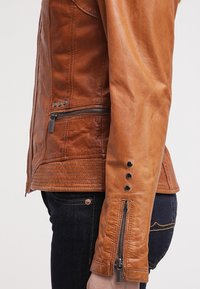 Giacca in pelle marrone con una finitura testurizzata, dotata di cerniere e dettagli metallici sulle maniche e un design slim e aderente.