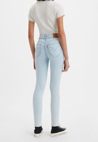 Jeans skinny de color azul claro con cintura alta, bolsillos traseros clásicos y un logo de parche de cuero marrón en la cintura. Llevados con zapatillas negras de caña alta.