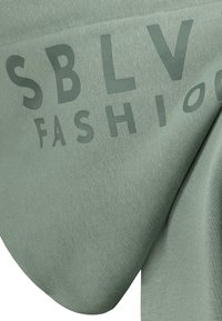 Grüner Stoff mit strukturierter Oberfläche, mit dem Text "SBLV FASHION" in einem leicht dunkleren Grünton bedruckt. Glatte Drapierung.
