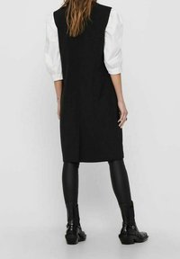 Czarna sukienka bez rękawów o dopasowanej sylwetce, zestawiona z białą bluzką z bufkami. Modelka ma na sobie czarne legginsy i botki za kostkę.