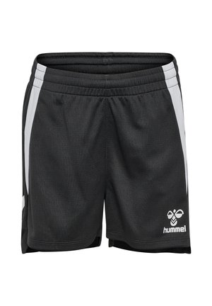 Ein junges Mädchen steht frontal da und trägt ein grau-schwarzes Sportoutfit mit weißen Socken und schwarzen Sneakers vor einem einfarbigen Hintergrund.