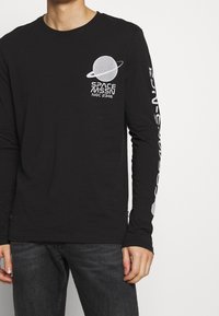 Svart långärmad t-shirt i bomull, med en texturerad planet och ringgrafik i vitt på bröstet och ärmen, samt tryckt text.