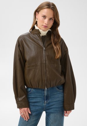 Jeune femme aux longs cheveux portant une veste zippée en cuir marron sur un col roulé blanc et un jean bleu sur un fond uni.