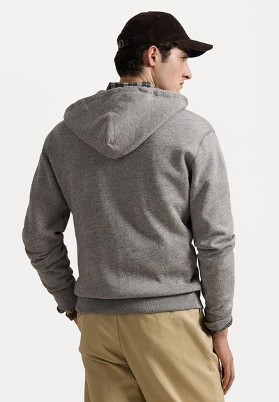 Hanorac gri dintr-un material texturat, cu buzunar tip kangur și glugă cu șnur, mânci cu model în vrac, purtat cu pantaloni bej și o șapcă maro.