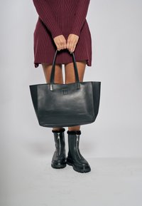 Borsa tote in pelle nera con due lunghi manici e un logo discreto. Abbinata a un abito bordeaux a coste e stivali neri con zip.