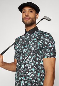 Kortermet polo skjorte med et svart blomster mønster med turkise, hvite og røde detaljer; kombinert med en svart caps og golfkølle.