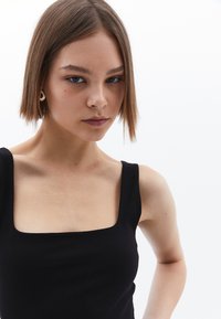 Haut noir à encolure carrée en tissu lisse, avec de larges bretelles. Modèle avec des cheveux courts, bijouterie minimale, sur fond blanc.