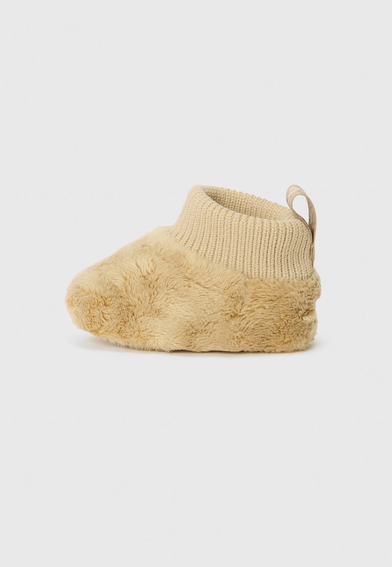 Chausson bébé beige doux avec corps en peluche, poignet côtelé en maille à la cheville et languette au talon sur un fond clair uni.