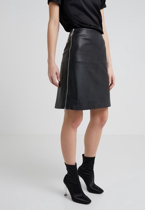 Femme portant une jupe en cuir noire avec une fermeture éclair sur le côté et des bottines noires à talons hauts, debout sur un fond uni.