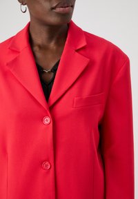 Red tailored blazer with a notch lapel, two buttons, and a chest pocket. Made from smooth fabric, featuring a sleek silhouette.