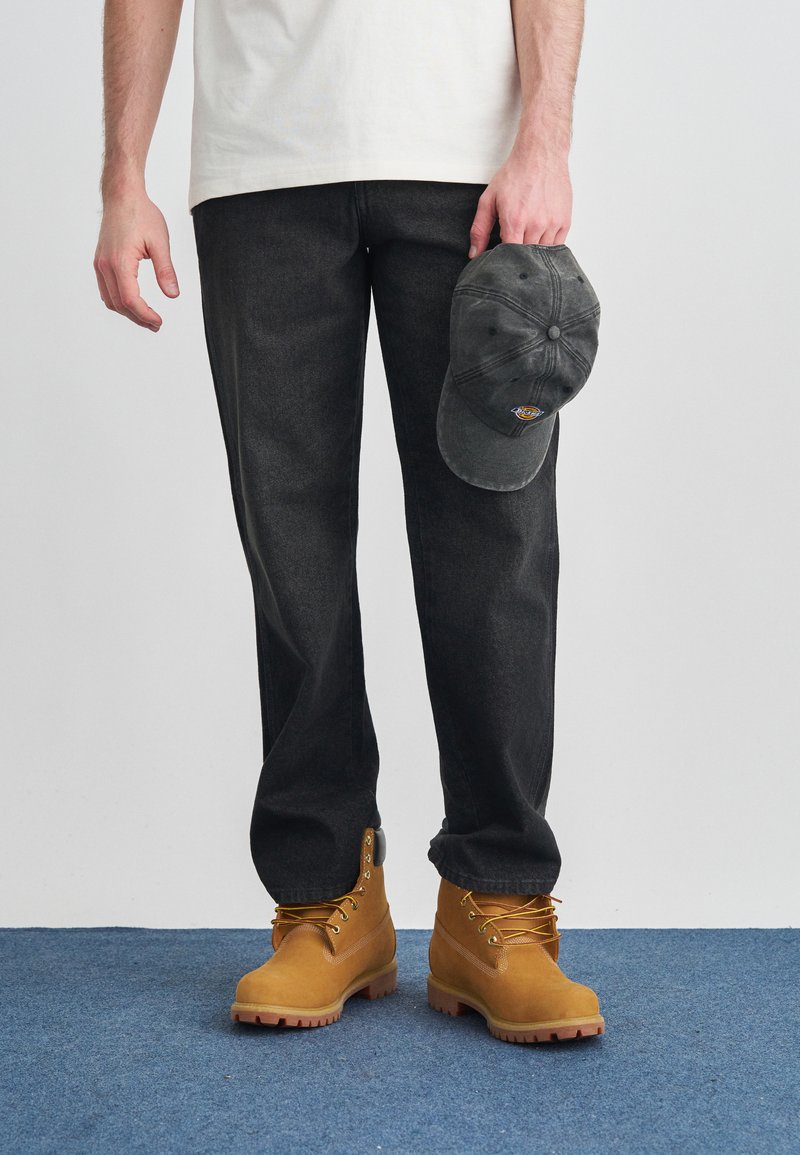 Jean gris foncé, bottes de travail marron clair avec lacets et semelles en caoutchouc, et une casquette de baseball grise tenue à la main. T-shirt blanc uni.