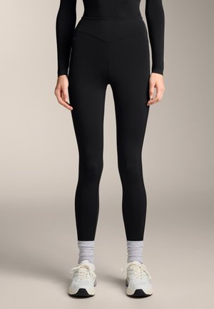 Leggings neri a vita alta realizzati in tessuto elasticizzato, con una superficie liscia e un design aderente, abbinati a sneakers bianche e calzini grigi.