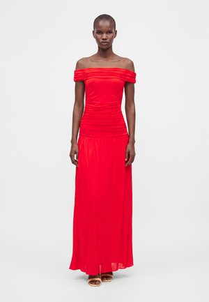 Femme portant une longue robe rouge vif bustier, debout devant un fond blanc uni avec une expression faciale neutre.