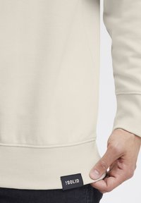 Sweatshirt beige clair en tissu doux avec poignets et ourlet côtelés. Présente une étiquette noire avec le texte « SOLID » en bas à droite.