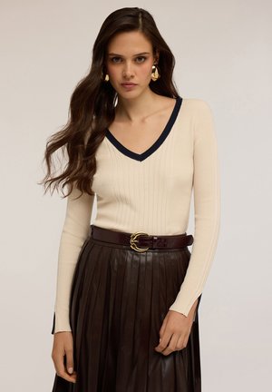 Donna con lunghi capelli castani che indossa un maglione a coste color crema con collo a V bordato di blu navy, orecchini a cerchio dorati e una gonna plissettata scura con cintura.