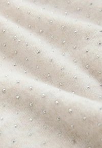 Tessuto beige chiaro con una morbida texture, arricchito da piccole paillettes argentate sparse come accento. Il design presenta un effetto luminoso e discreto.