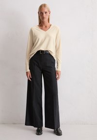 Beżowy sweter w serek nałożony na szerokie ciemnoszare spodnie utrzymywane w pasie czarnym paskiem, w zestawieniu z czarnymi butami. Tekstura wydaje się miękka.