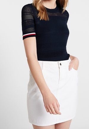 Femme portant un haut en maille bleu marine à manches courtes avec des rayures rouges, blanches et bleues sur la manche et une jupe en jean blanche avec des poches devant.