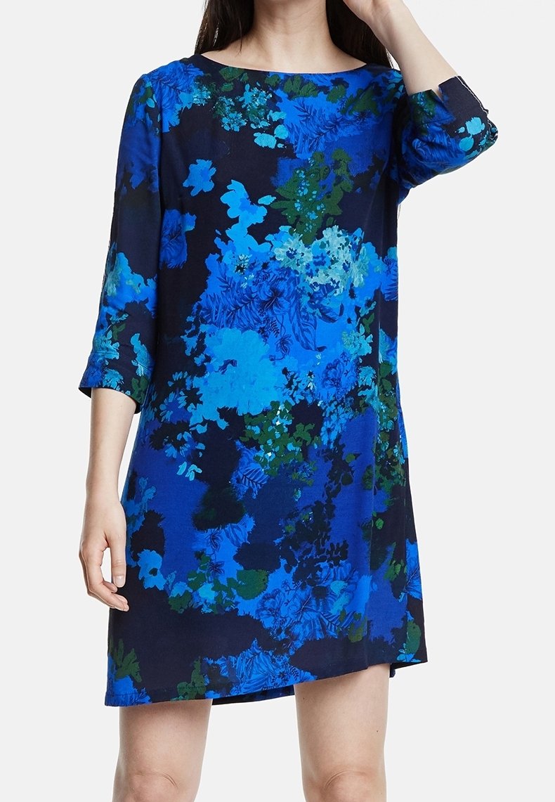 Femme portant une robe longueur genou avec des manches 3/4, présentant un motif floral bleu et vert vif sur un fond sombre.