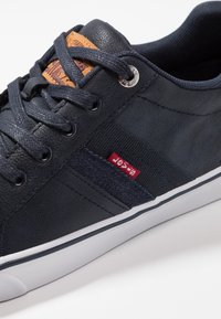 Baskets bleu marine avec une tige en cuir texturé, une bande latérale en tissu contrastant et une étiquette en cuir marron. Elles disposent de lacets plats et d'une semelle en caoutchouc.