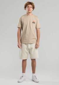 Beige cotton t-shirt with "Peace of Mind" graphic and light-colored shorts. The model wears white sneakers. Simple, casual design.