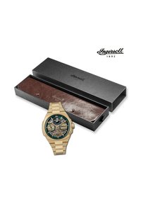 Gouden polshorloge met een groene wijzerplaat, zichtbare tandwielen en twee sub-dials. Gepresenteerd in een zwarte doos met een binnenkant van bruin leer.