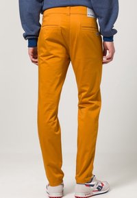 Gele katoenen broek met een slanke pasvorm, voorzien van achterzakken en een label op de tailleband, gecombineerd met witte en rode sneakers.