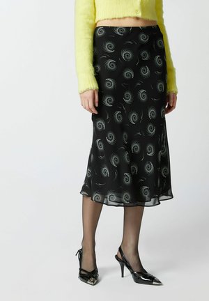 Falda negra de slip con patrón plateado en espiral, hecha de un tejido ligero; combinada con un suéter corto amarillo y esponjoso y unos tacones negros brillantes.