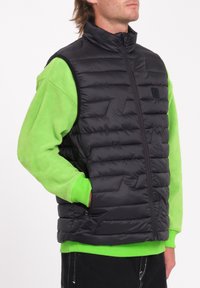 Gilet imbottito nero con colletto alto, chiusura con zip e applicazione del logo, indossato sopra una felpa in pile verde brillante. Tessuto testurizzato e leggero.
