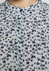 Blumenbluse aus leichtem Stoff, hellblauer Hintergrund mit navyblauen Blumen und schwarzen Stielen, mit Stehkragen und einem schwarzen Knopf.