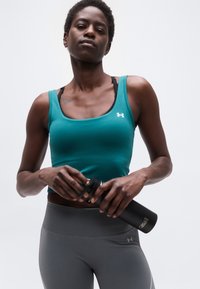 Ευτραφής άτομο που φοράει τιρκουάζ αθλητικό τοπ και γκρι κολάν, κρατώντας μαύρο μπουκάλι νερού Casall, με φόντο απλό λευκό.