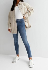 Beige knappljacka över en vit topp, i kombination med smala blå jeans och vita sneakers. Anmärkningsvärda detaljer: fransat fåll på jeansen, bröstficka på jackan.