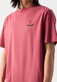 Camiseta de algodón roja con mangas cortas, que presenta un pequeño diseño de globo y texto en negro y dorado en la parte superior izquierda. Textura suave.