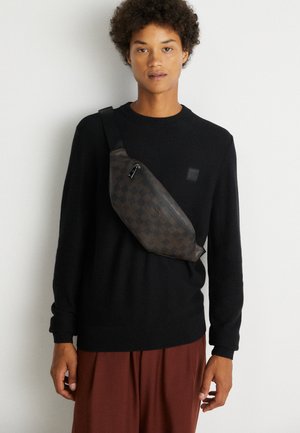 Ung person iført sort sweater og brune bukser med en mørk ternet crossbodytaske mod en ensfarvet hvid baggrund.