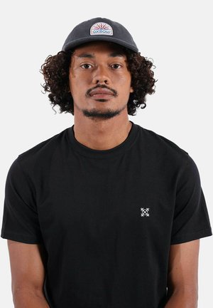Homme aux cheveux bouclés portant une casquette de baseball sombre et un t-shirt noir uni avec un petit logo blanc brodé sur la poitrine.