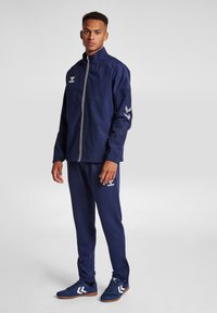 Marinfärgad träningsoverall med vit dragkedja och logotyp. Jackan har marinfärgad panel och texturerade ärmar; byxorna har sidodragkedjor och matchande sneakers.