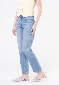 Mujer vestida con un chaleco amarillo claro, jeans azules y zapatos de tacón tipo slingback en rosa pálido, de pie contra un fondo blanco.
