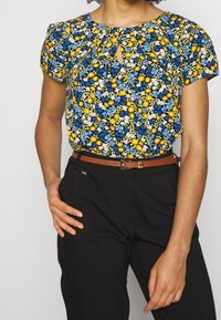 Blusa floreale con una stampa blu, gialla e bianca, caratterizzata da maniche corte e scollatura a chiave, abbinata a pantaloni neri e una cintura marrone.