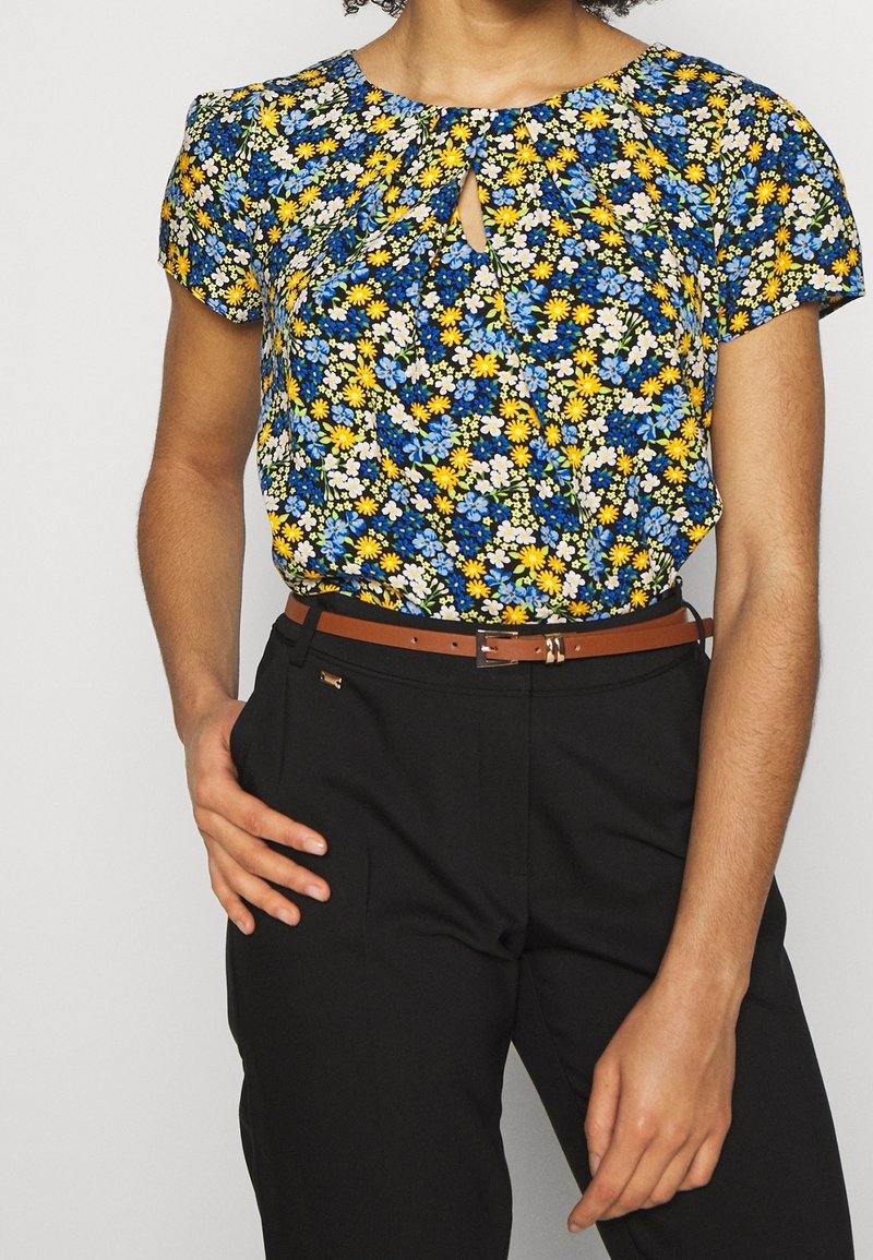 Blusa floreale con una stampa blu, gialla e bianca, caratterizzata da maniche corte e scollatura a chiave, abbinata a pantaloni neri e una cintura marrone.