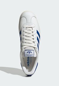 Bijela tenisica s plavim prugama, izrađena od kože i mrežastog materijala, s okruglim vrhom, ravnim vezicama i Adidas logom na jeziku.