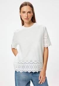 Camiseta de algodón blanca con mangas cortas, que presenta un dobladillo ondulado y un intrincado diseño de recortes a lo largo de los bordes. Ajuste casual y textura suave.