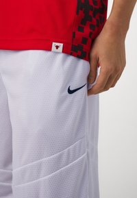 Ręka spoczywająca obok białych siateczkowych spodenek Nike z granatowym symbolem Swoosh, czerwonej sportowej koszulki z czarnym pikselowym wzorem i metką z logo Chicago Bulls.