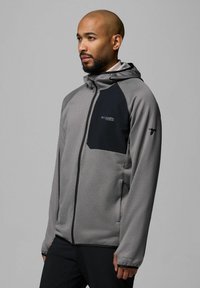 Lekka szara kurtka o teksturowanej powierzchni, z czarnymi akcentami, kapturem i zamkiem, z logo na klatce piersiowej i rękawie.