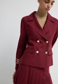 Burgundy double-breasted blazer with large gold buttons, tailored fit, and notched lapels, paired with a pleated skirt. Smooth fabric texture.